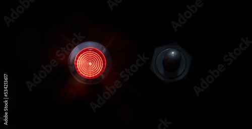 Switched ON. Red indicator or ON lamp with a toggle switch next to it. Red status lamp with a retro toggle switch on black panel. Shallow depth of field.
