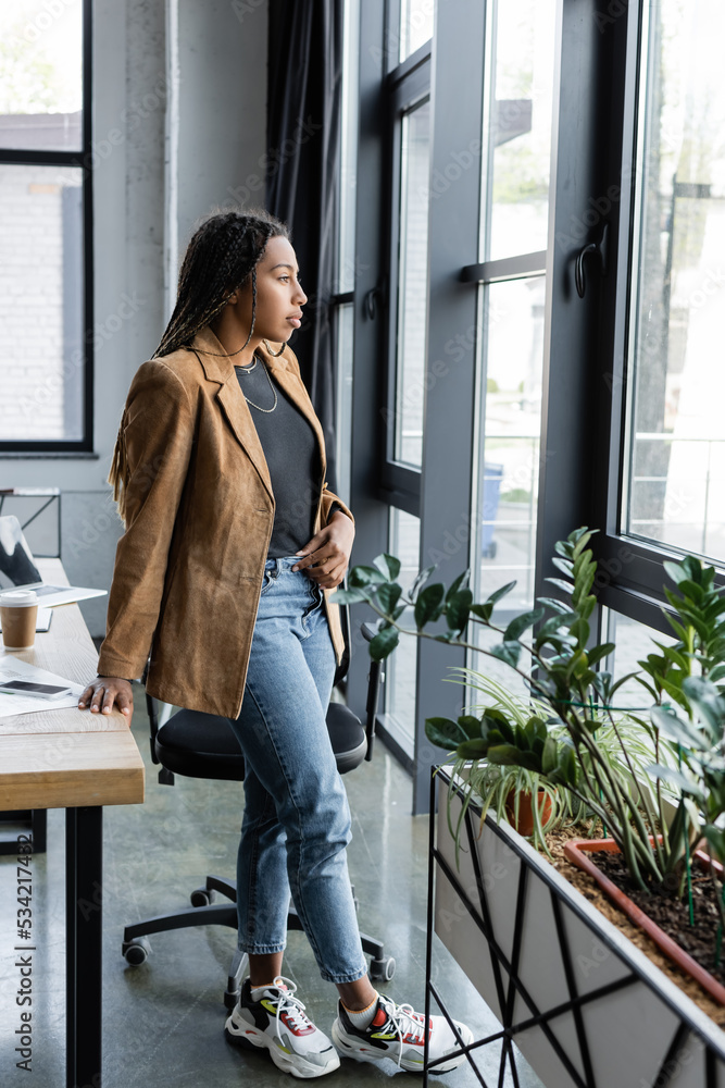Side view of african american businesswoman in jacket and jeans looking at window in office. Side view of african american businesswoman in jacket and jeans looking at window in office.