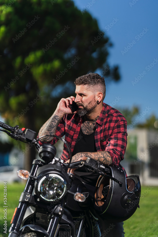 Chico con camisa a cuadros roja posando con telefono movil en su motocicleta