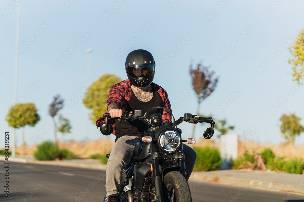 Chico joven tatuado con camisa a cuadros roja montando motocicleta por carretera solitaria al atardecer Chico joven tatuado con camisa a cuadros roja montando motocicleta por carretera solitaria al atardecer