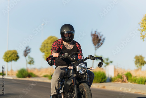 Chico joven tatuado con camisa a cuadros roja montando motocicleta por carretera solitaria al atardecer © MiguelAngelJunquera