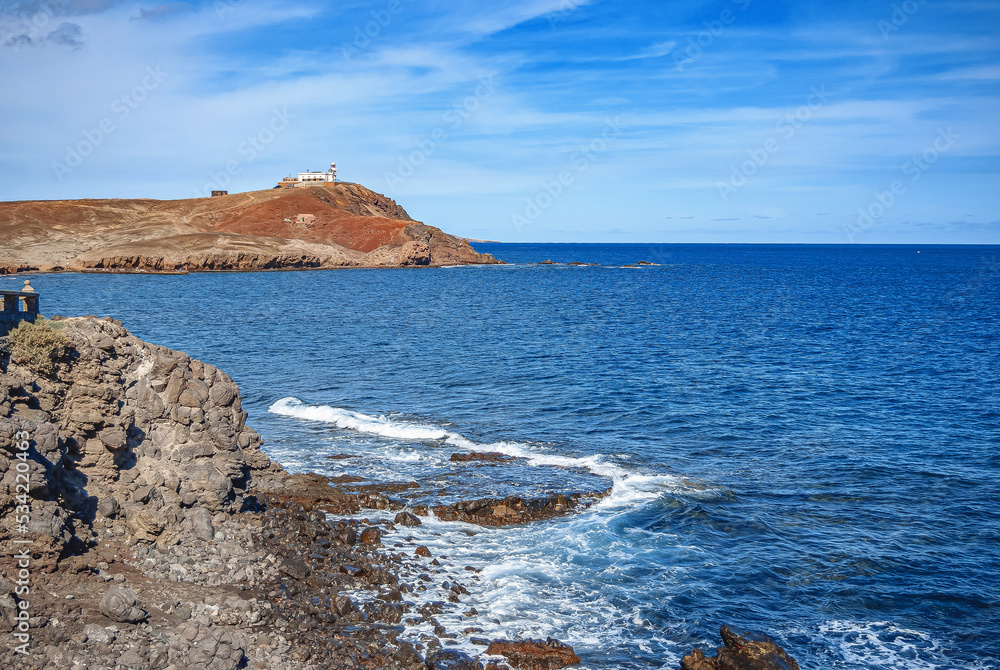 A view of the ocean and a small lighthouse at the entrance pier of the island. Calm blue sea and sky, stone shore.
