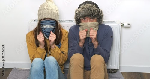 Funny people at home apartment freezing from winter cold. Woman and man sitting near radiator in warm hats, pull scarves over noses. Shaking, shivering in chilly room. Poor heating in house concept photo