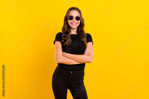 Photo of adorable confident young lady dressed black clothes dark eyewear arms crossed smiling isolated yellow color background