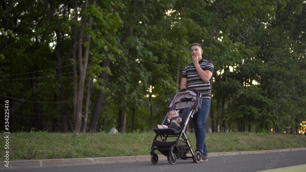 Obraz premium A man rolls a baby around the park and eats a burger. Young father with his son for a walk.