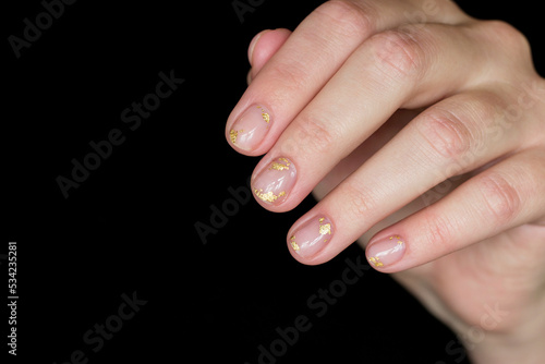 gentle aesthetic trendy manicure on short nails with transparent gel polish with decor pieces of gold foil close-up on fingernails on a black background