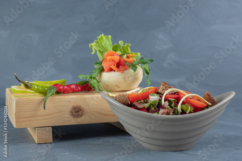 Gray salad bowl next to a bundle of vegetables on a wooden board on marble background