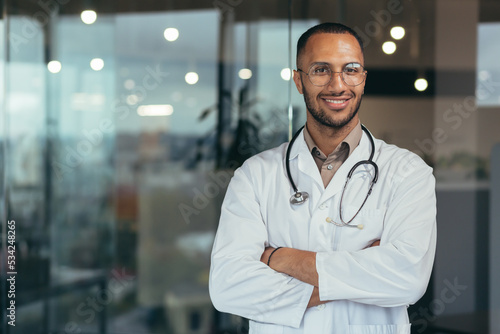 Photos Portrait of young happy and smiling doctor, man in medical coat and stethoscope smiling and looking at camera, Arab doctor with crossed arms working inside modern clinic office