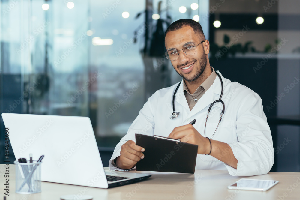 Portrait of successful male doctor, hispanic smiling and looking at camera, doctor in medical gown working inside modern clinic office, using laptop for work Portrait of successful male doctor, hispanic smiling and looking at camera, doctor in medical gown working inside modern clinic office, using laptop for work