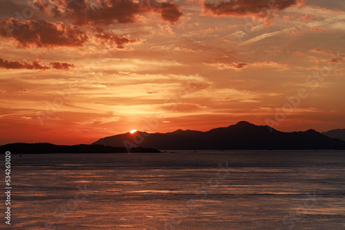 Offshore view of sun setting behind mountains as sky glows orange