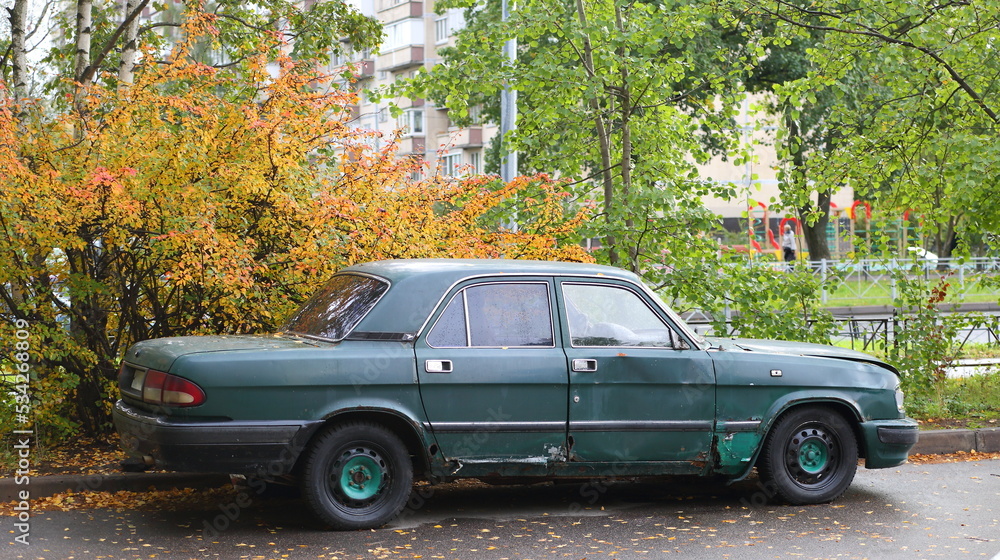 An old rusty green car is parked near the autumn bush, Podvoysky Street, St. Petersburg, Russia, September 2022