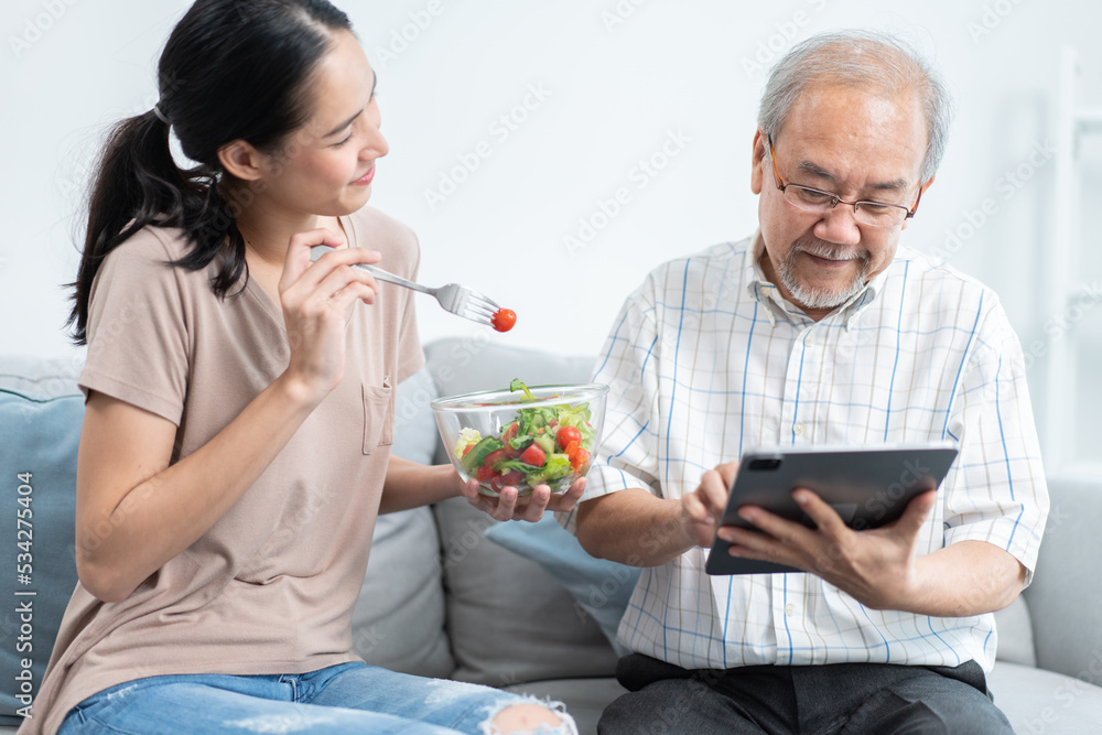 Young Asian daughter serving and feeding food to senior father while he using and playing on digital tablet in the living room. family love and support to elderly people. 