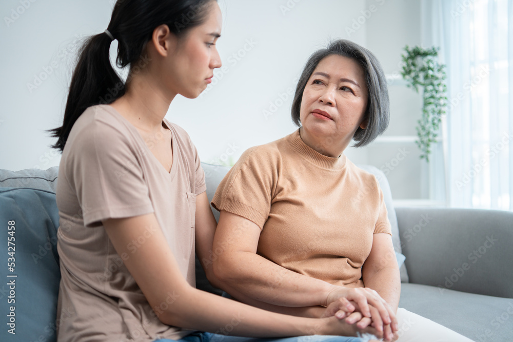 Asian senior mother consoling unhappy daughter, holding hands and support to young depressed woman. Mom talking and giving advice to solve problem sitting on couch at home. 