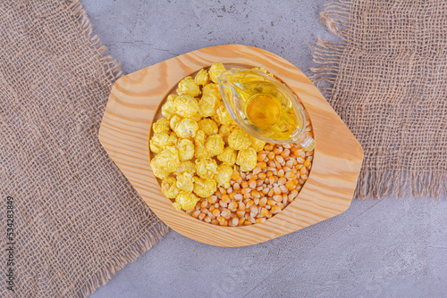 Wooden plate on two pieces of fabric, with a pile of kernel, a portion of caramel popcorn and a slamm glass of oil on marble background