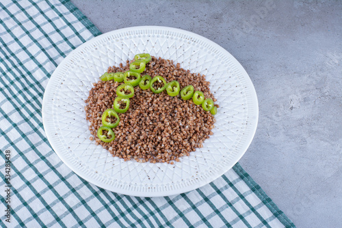 Pile of buckwheat topped with pepper slices on an ornate plate on a towel on marble background