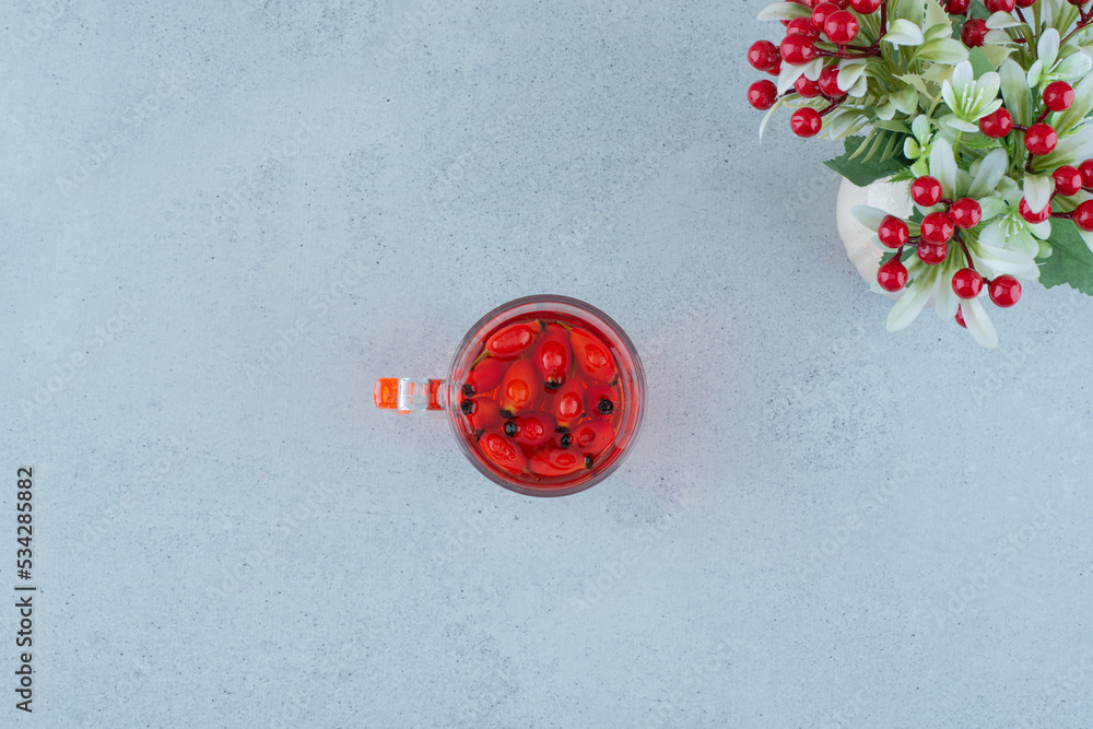 Glass of juice with rosehips and artificial flowers on blue surface