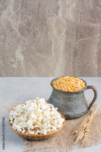 A small weaved basket of popcorn, a jug full of corn grains and stalks of wheat lined up on a piece of cloth on marble background