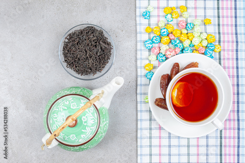 A composition of teapot, a small bowl of tea leaves and a cup of tea on a towel with scattered popcorn candy on marble background