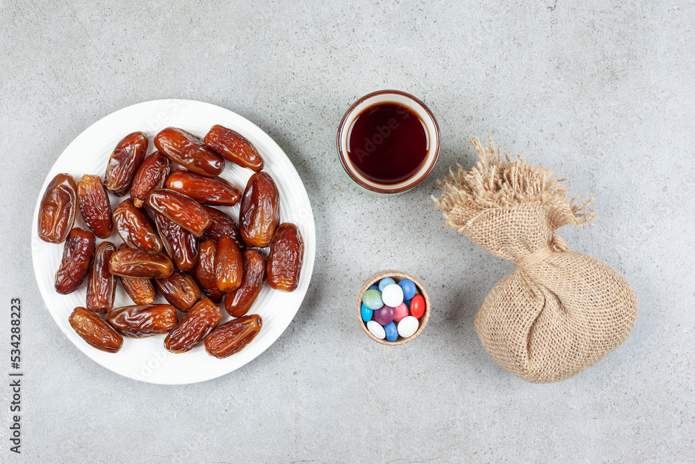 A platter of dates, a cup of tea, a sack and a small bowl of candies on marble background A platter of dates, a cup of tea, a sack and a small bowl of candies on marble background