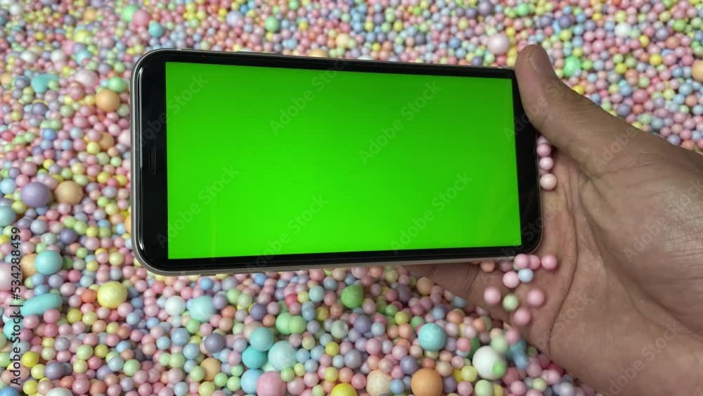 Chroma key green screen on smartphone landscape in male hand with colourful pastel rainbow foam ball beads in background Chroma key green screen on smartphone landscape in male hand with colourful pastel rainbow foam ball beads in background