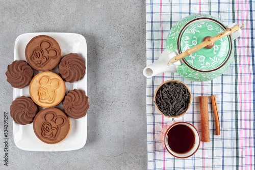 Cookies arranged on a plate next to ornate teapot, cinnamon cuts,cup of tea and a bowl of tea leaves on marble background