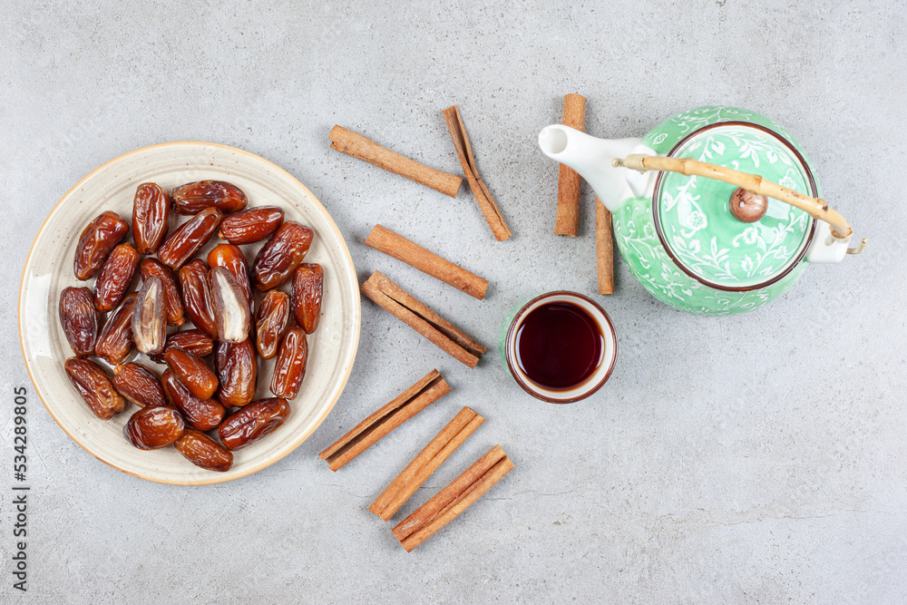 Fototapeta premium A cup of tea, scattered cinnamon cuts and an ornate teapot next to a plate of fresh dates on marble background