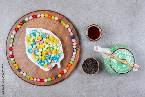 Teapot and a bowl of tea leaves next to a cup to tea, with candies arranged on a wooden board on marble background