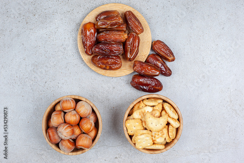 An assortment of dates on wooden board and nuts and biscuits in bowls on marble background