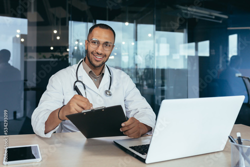 African american doctor portrait, man working inside modern clinic office at table using laptop, doctor in medical coat and stethoscope smiling and looking at camera.