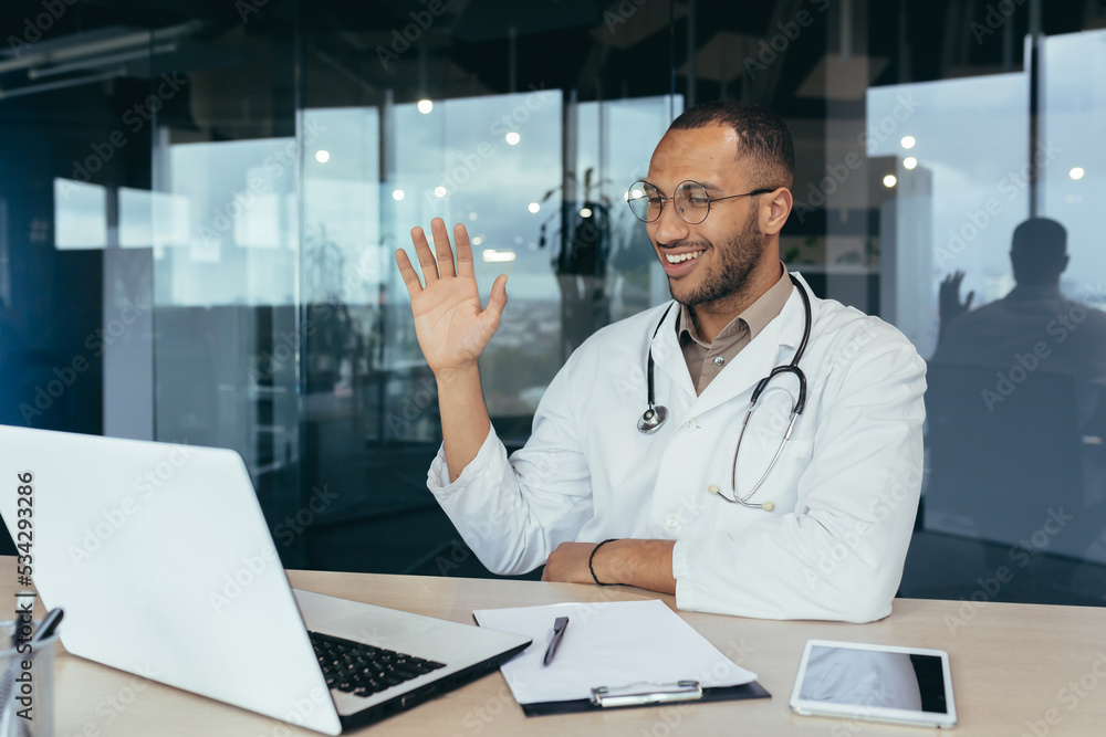 Happy hispanic doctor smiling and talking on video call man in white coat and stethoscope using laptop for remote online consultation smiling and waving hello. Happy hispanic doctor smiling and talking on video call man in white coat and stethoscope using laptop for remote online consultation smiling and waving hello.