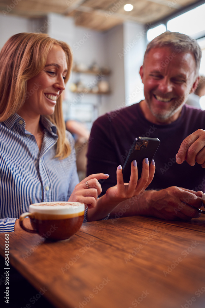 Couple Sitting At Table In Coffee Shop Looking At Mobile Phone Together