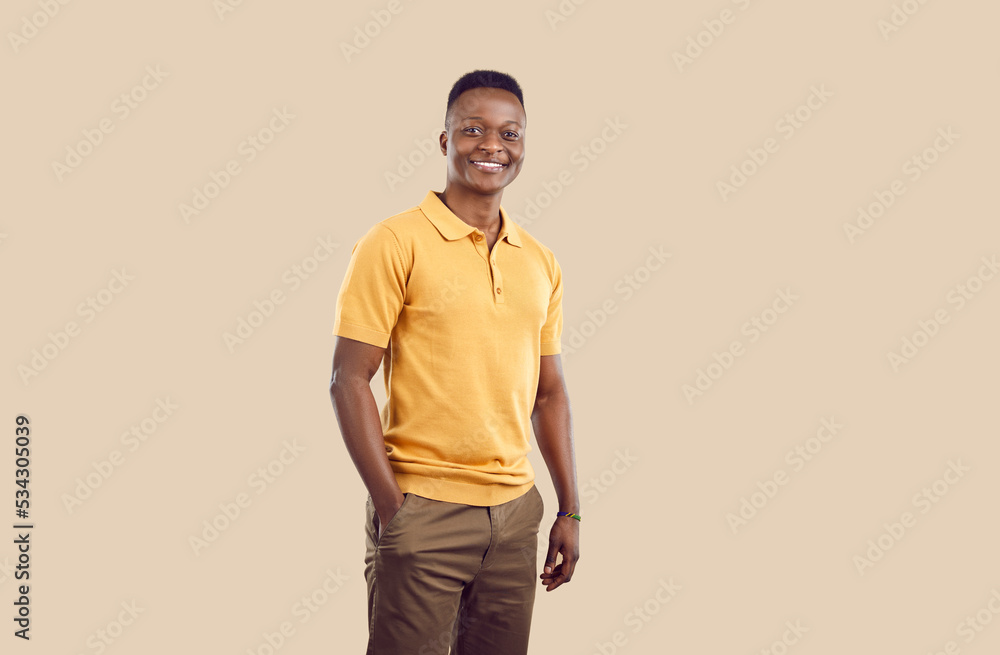 Fototapeta premium Young smiling African American man millennial with short hair posing with one hand in pocket pants and confidently looking at camera dressed in casual clothes stands on beige background. Copy space
