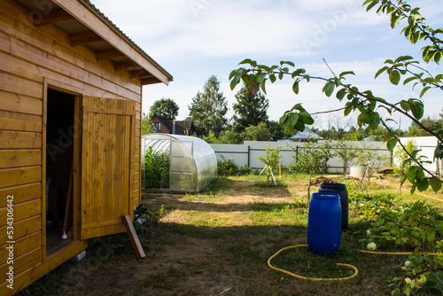 A wooden shed and large blue plastic water barrel stands in the garden among trees and green grass on a sunny summer day in the Moscow region Russia