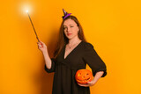 Young woman in black dress and small purple witch hat holding orange Jack o lantern pumpkin and looking at camera. Wizard girl holding luminous magic wand or glowing magic stick on yellow background.