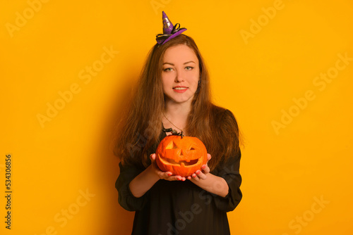 Happy young smiling woman holding Halloween pumpkin or Jack o lantern pumpkin with toy spider and looking at camera on a yellow background.Wizard girl or witch girl in black dress with a purple hat.