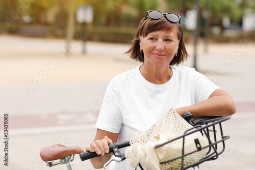 Portrait of active middle ages woman on bicycle on sunny day. Mature lady in white t shirt is riding city bike in park. Leisure and lifestyle concept, smiling female cycling with her bike in summer