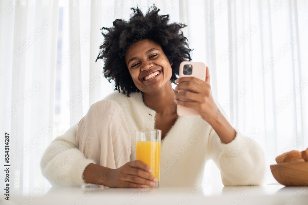 Portrait of young woman working from home holding glass of juice. Happy female model drinking juice, looking at camera, holding smartphone