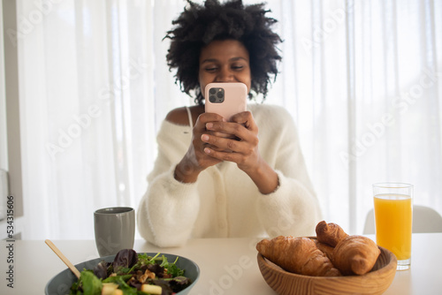 Portrait of young woman working from home holding phone smiling. Happy female model typing on smartphone, sitting at table with breakfast. Portrait, remote work concept.
