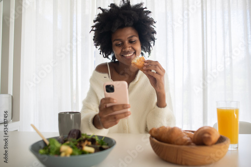 Portrait of young woman working from home holding croissant. Happy female model having bite of croissant, smiling, sitting at table, holding smartphone. Portrait, remote work concept.