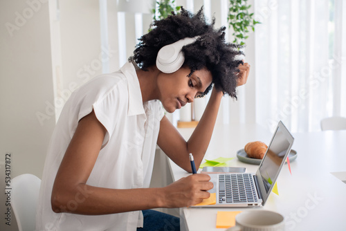 Concentrated woman in headphones writing on sticky paper. Focused African American woman sitting at computer desk and studying online during self-isolation. Online education, pandemic concept