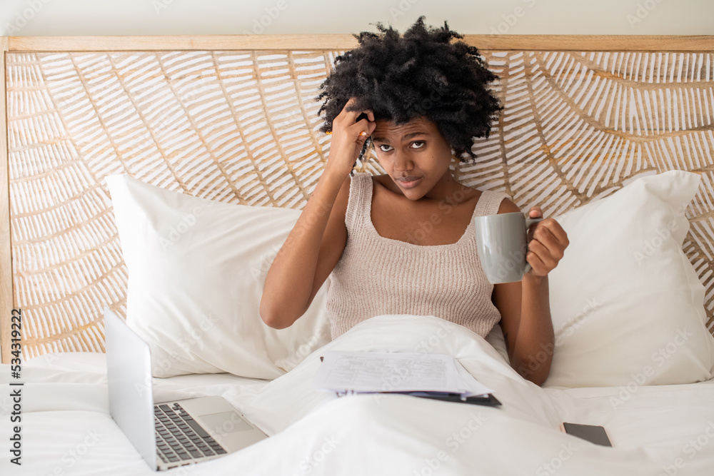 Pensive businesswoman working in bed after waking up. Vertical shot of African American woman drinking tea, thinking, working with papers, using laptop. Home office, paperwork, freelance concept Pensive businesswoman working in bed after waking up. Vertical shot of African American woman drinking tea, thinking, working with papers, using laptop. Home office, paperwork, freelance concept