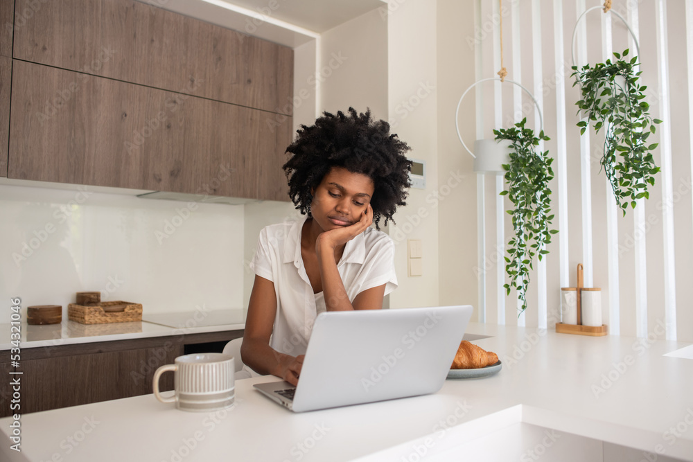 Sad African-American woman working on laptop in morning. Pensive woman sitting at table in kitchen and working from home while having breakfast. Home office, business, technology concept