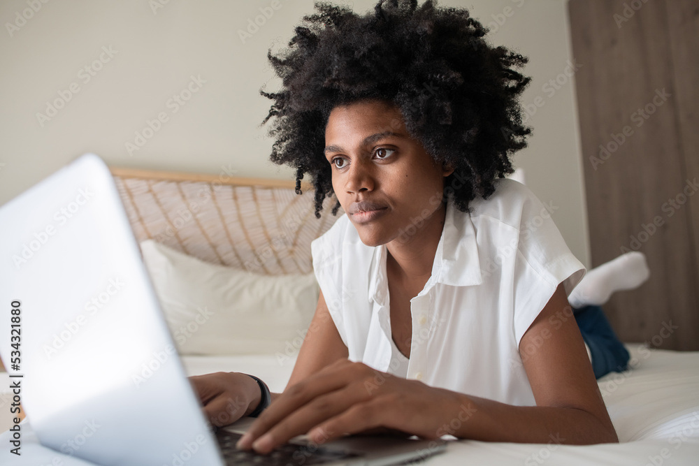 Focused woman lying on bed and looking at laptop screen. Horizontal shot of African American freelancer working from home, analyzing information. Home office, business, occupation concept Focused woman lying on bed and looking at laptop screen. Horizontal shot of African American freelancer working from home, analyzing information. Home office, business, occupation concept