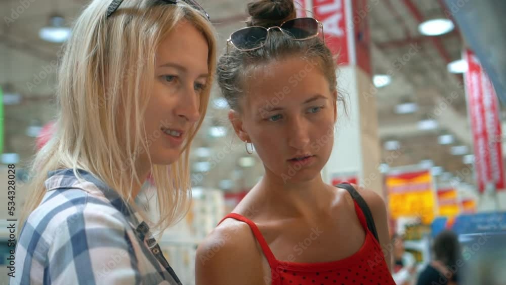 Two young Caucasian women choose glasses in dishes department in supermarket. Girls are happy, laughing and happy enjoying shopping in store. Happy friends or sisters, customers in store. Two young Caucasian women choose glasses in dishes department in supermarket. Girls are happy, laughing and happy enjoying shopping in store. Happy friends or sisters, customers in store.
