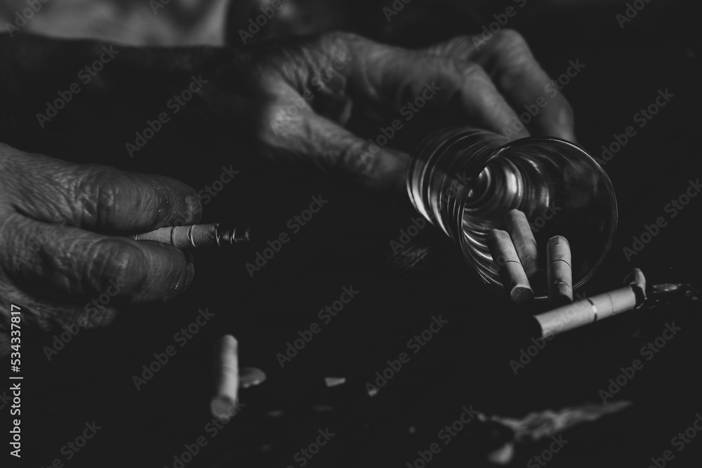 Cigarettes a glass and an empty bottle and the hands of an old woman on a burnt black table, female smoking and alcoholism