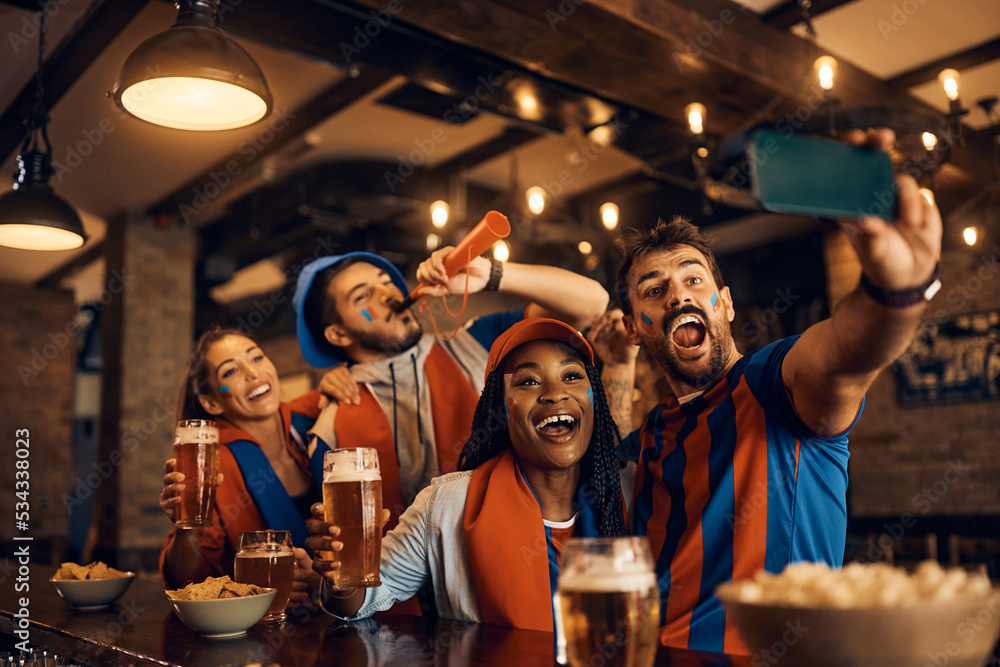 Cheerful sports fans taking selfie while celebrating their team's victory in pub.