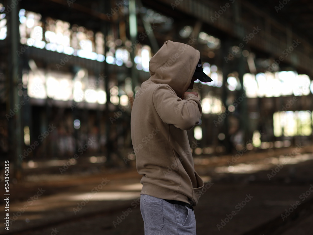 a young rapper guy in a hood and cap black jacket in an abandoned old building - obrazy, fototapety, plakaty 