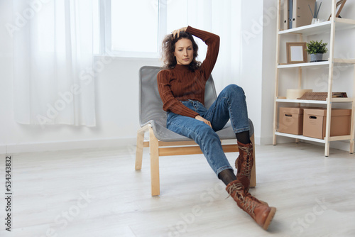 Upset pensive young frustrated adorable curly woman think about difficult life decision about divorce reclines on hand touch temple sitting in chair at home. Social media online communication concept