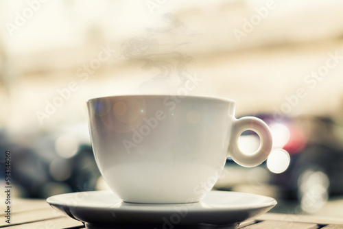 Ceramic white cup with hot coffee stands on wooden table of street cafe against blurred road traffic