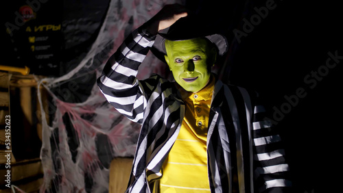 Halloween party, night, twilight, in the rays of light, a man with a terrible make-up, with a green face and a hat lifts his hat, reveals his tongue, laughs eerily and runs away. High quality photo